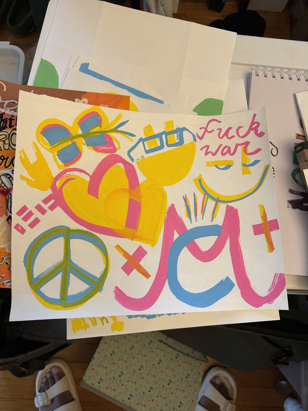 Painted sheet with a peace sign, a butterfly, overlapping hearts, a face with glasses, a large M in pink and blue, and the words fuck war in pink script